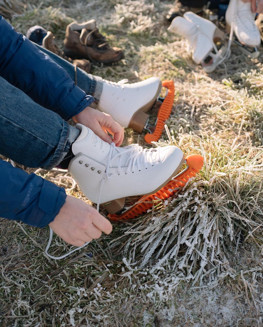 Woman ties laces on ice skates outdoors on frosted grass. Additional skates and shoes are in the background, winter activity.