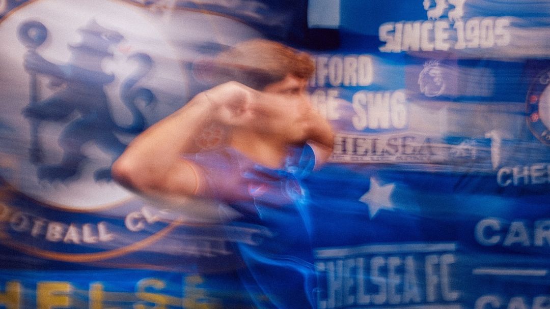 Blurred motion shot of a soccer fan in blue jersey at a stadium, with Chelsea FC logo and text 'Since 1905' visible in the background.