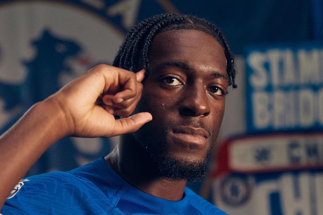 Close-up portrait of a Black man with braided hair wearing a blue shirt, posing in front of Chelsea soccer team branding.
