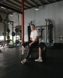 A man with prosthetics seated on a black box in a gym, with gym equipment in the background.