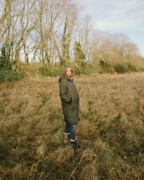 Woman in an olive green puffer coat standing in a dry grassy field before a line of bare trees.
