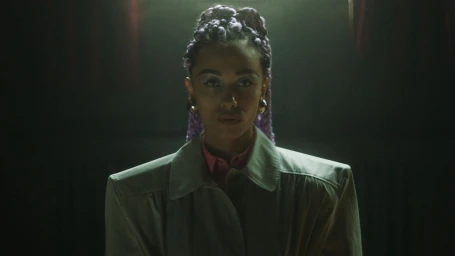 Portrait of a woman with braided purple hair wearing a structured grey coat and pink shirt against a dark background