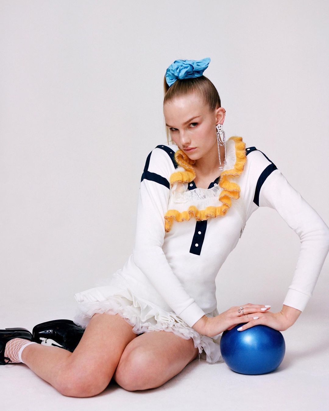 Young woman in a white dress, black shoes, and blue scrunchie sitting on a white surface and posing with a blue ball.