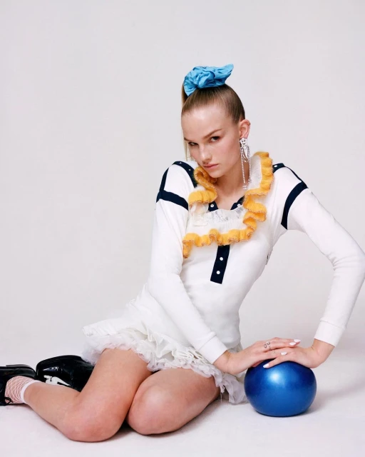 Young woman in a white dress, black shoes, and blue scrunchie sitting on a white surface and posing with a blue ball.