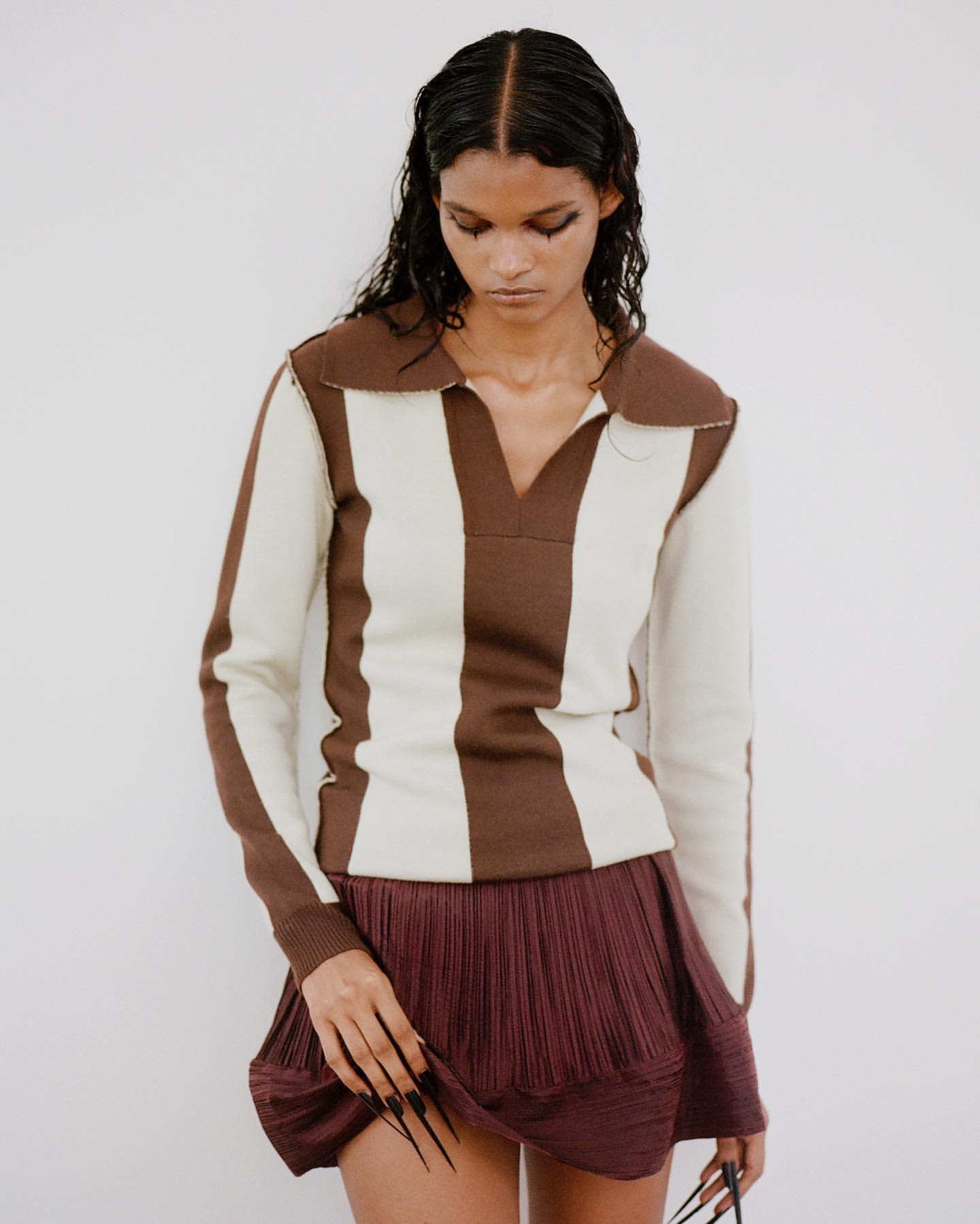 Woman with wet hair poses in studio wearing brown and white striped top and burgundy skirt, showcasing long stiletto fingernails.