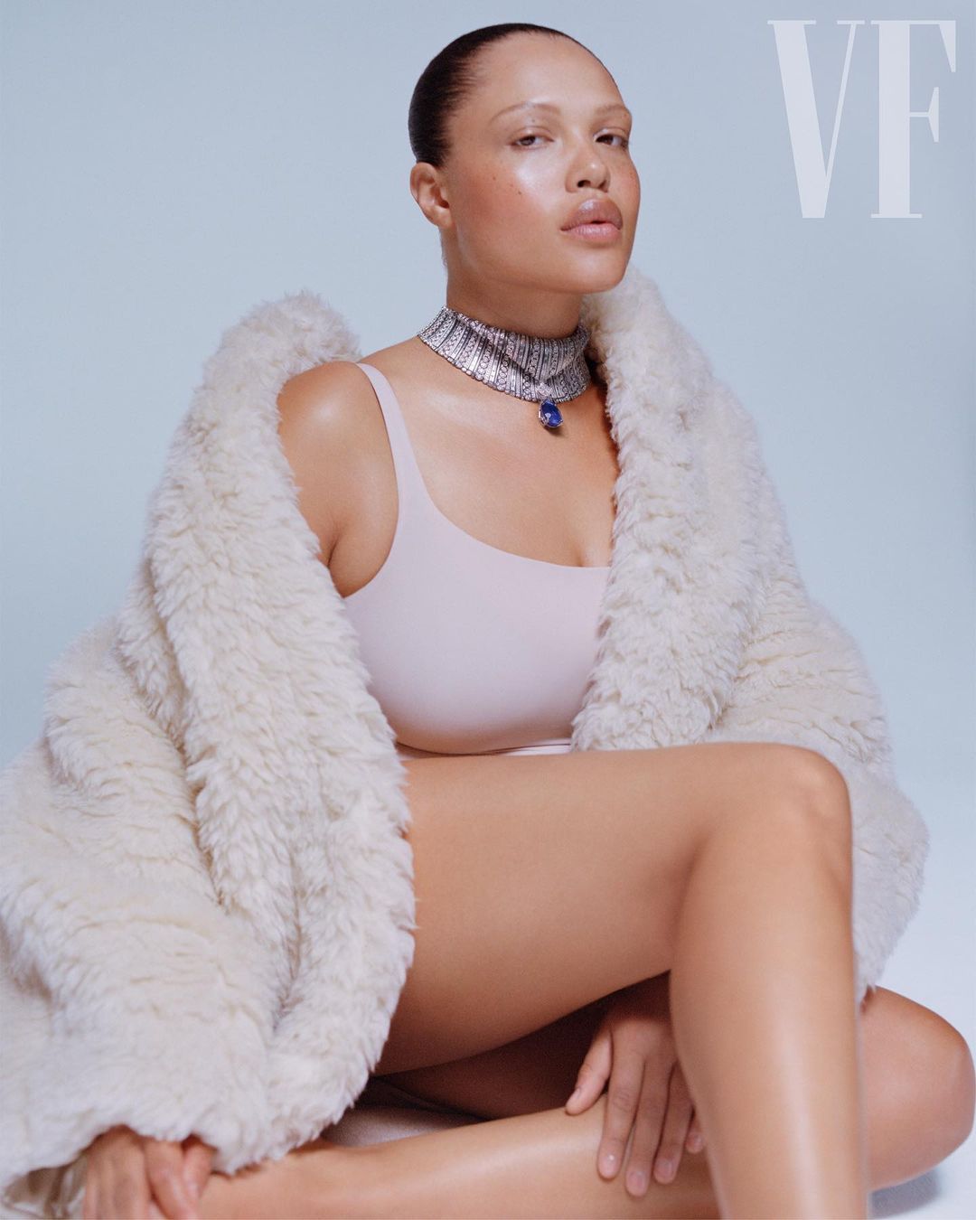 Woman poses in a fur coat and jewelry against a light blue backdrop, showcasing fashion and portrait photography.