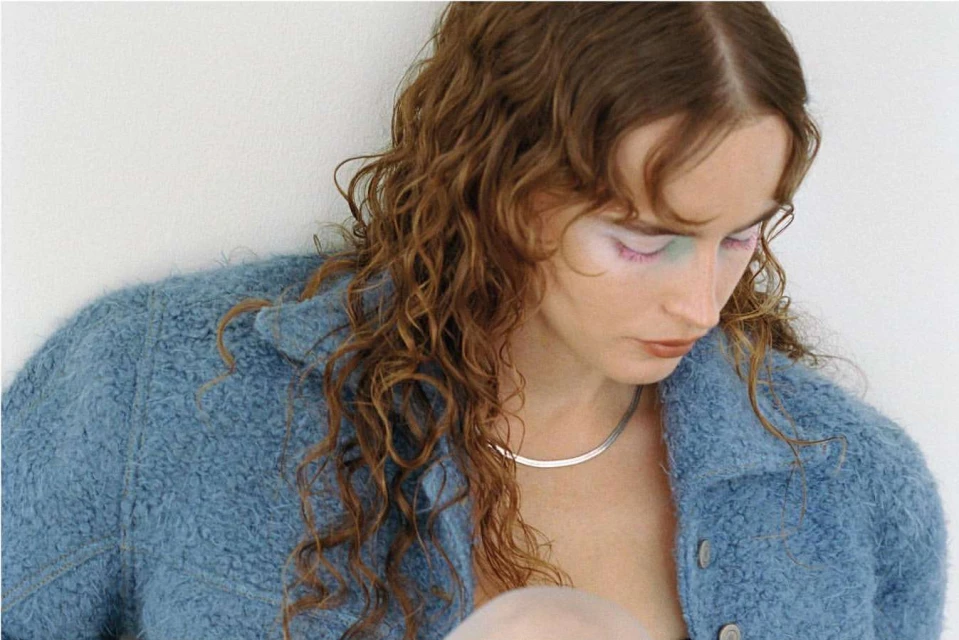 Portrait of a young woman with wavy hair and blue jacket looking down against a white background in a fashion style pose.