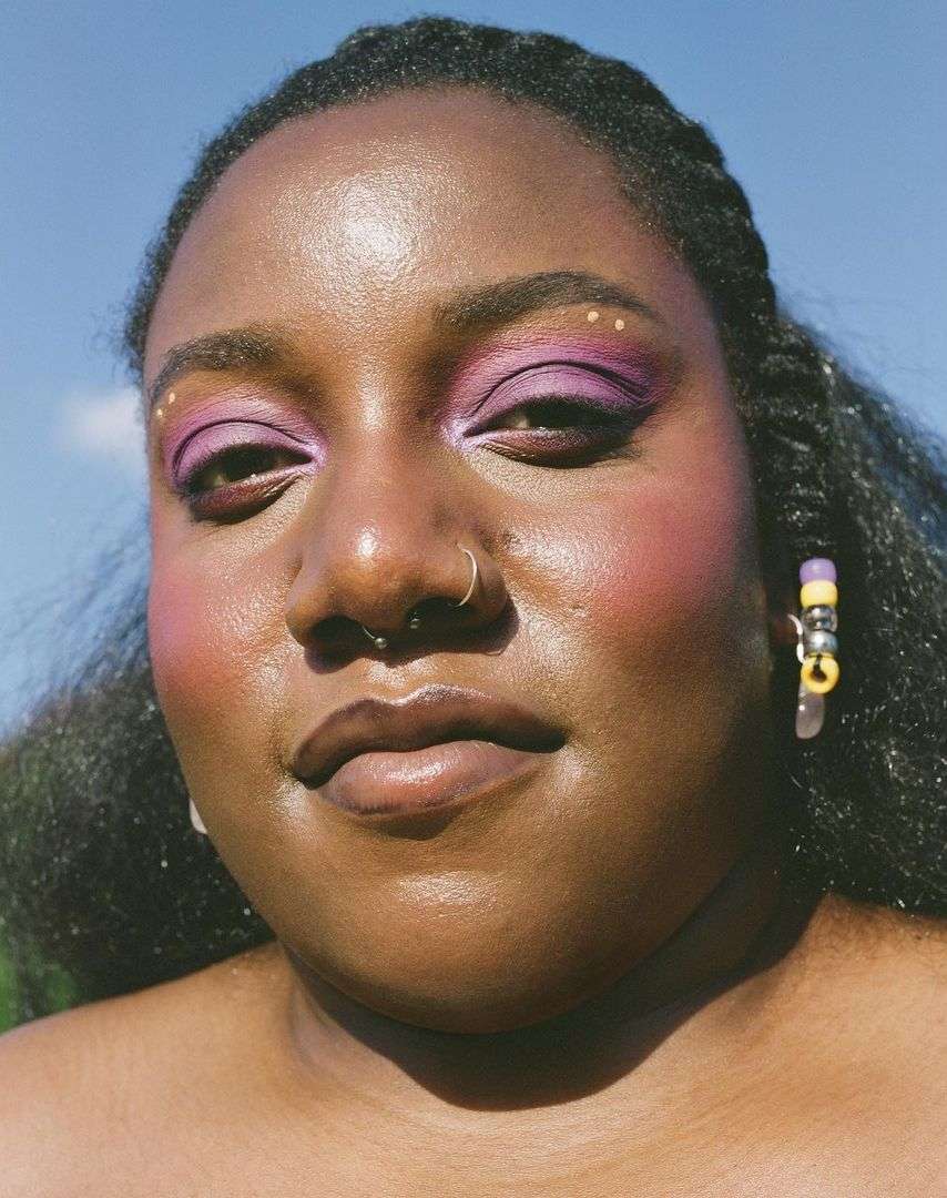 Close-up portrait of a smiling Black woman with violet and pink makeup, hoop earrings, and braided hair set against a bright blue sky.