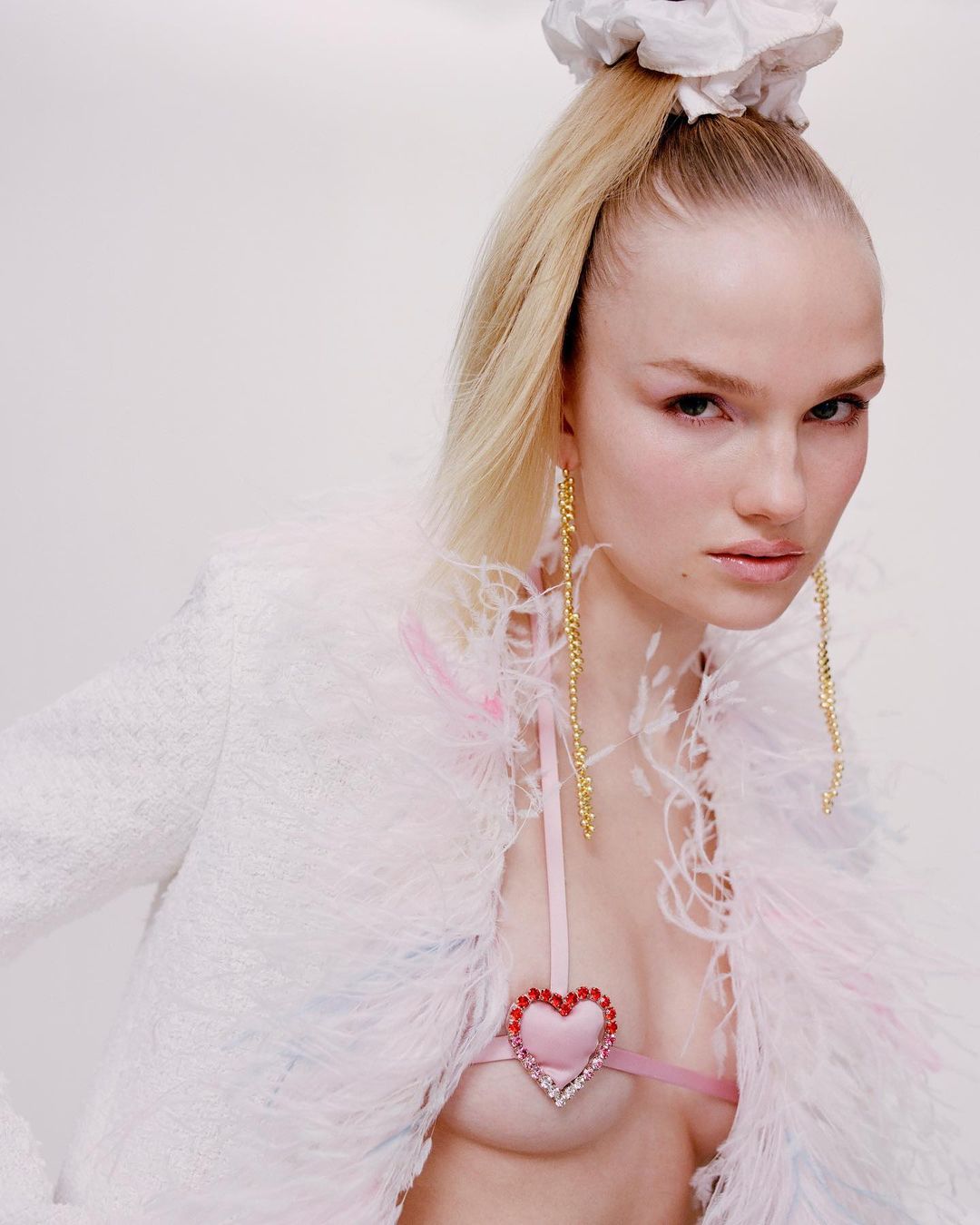 Blonde woman in a textured white jacket with feather trim and a heart-shaped bra, styled with a high ponytail and long earrings, against a white backdrop.