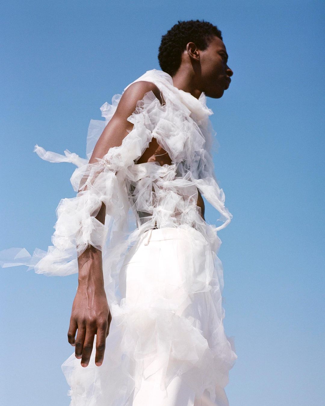 A dark-skinned male fashion model poses in a frilly white garment against a clear blue sky, showcasing unique sartorial design.