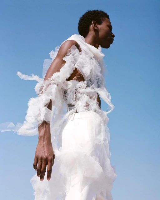 A dark-skinned male fashion model poses in a frilly white garment against a clear blue sky, showcasing unique sartorial design.