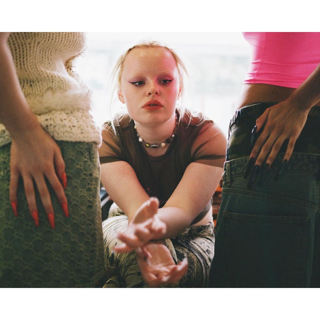 Portrait of a young woman with fair skin, dark eyeliner, and light lipstick, posing with two other women in an indoor setting.