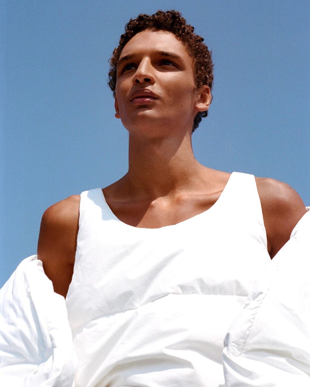 Portrait of a dark-skinned male model wearing a white tank top and puffer jacket against a clear blue sky, looking up in bright daylight.
