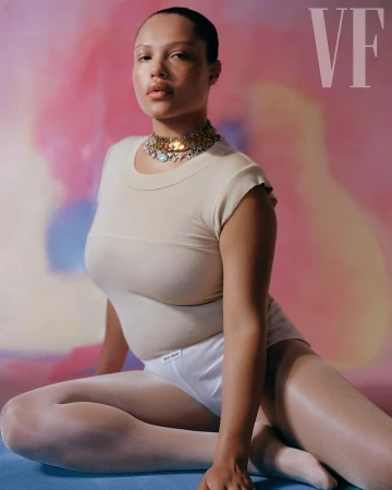 Fashion shot of a plus-size woman in a light beige top and white bottoms sitting cross-legged against a soft, colorful background.