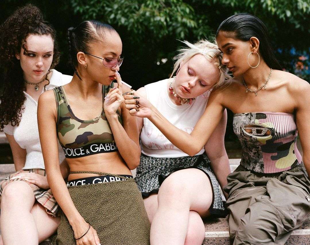 Four young women in trendy casual fashion sit outside in a park setting with a green leafy tree in the background, enjoying daylight.