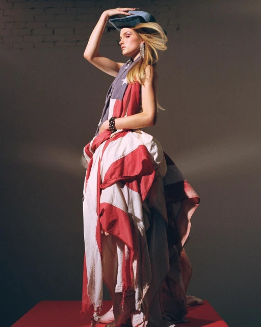 A woman in a patriotic fashion dress and hat stands on a red platform, creating a striking studio portrait with a gray backdrop.