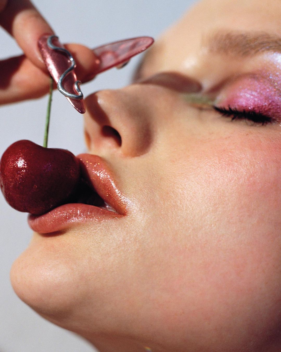 Close-up of a woman's face with glossy makeup and long fingernails, holding a cherry to her lips, bright lighting.