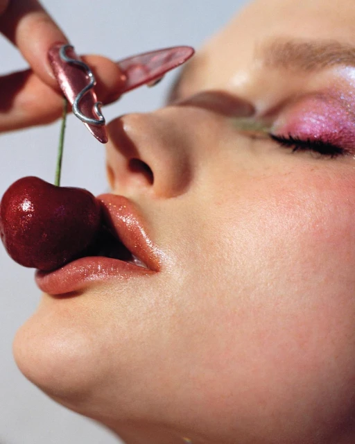 Close-up of a woman's face with glossy makeup and long fingernails, holding a cherry to her lips, bright lighting.