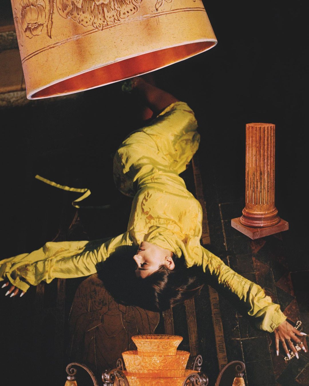 Woman in yellow dress poses beneath a large lamp, lying on an ornate floor with vintage column and decor for a fashion editorial.