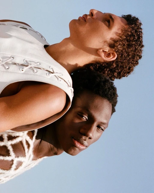 Two fashion models pose in white textured tops against a clear sky in a medium shot, focusing on skin tones and textured fabric details.