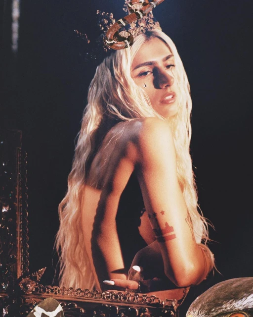 Portrait of a fair-skinned adult female with long blond wavy hair, wearing a jeweled crown and snake, looking over shoulder in dark studio.