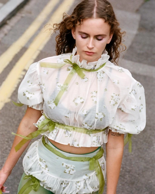 Young adult female model wears a floral-patterned ruffle blouse and skirt with green ribbons, posing outside on a city street with curly hair.