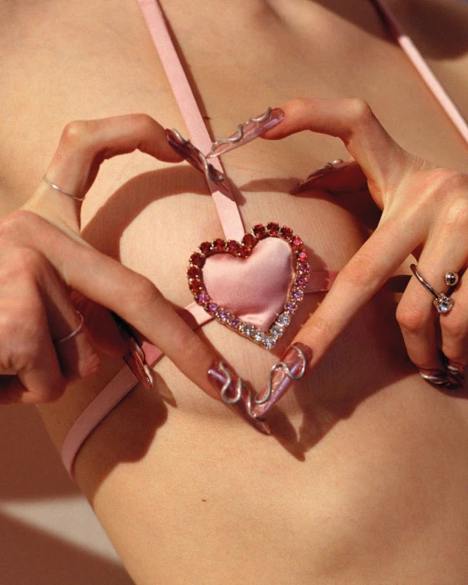 Close-up of woman's torso with embellished heart pendant on pale pink top, finger jewelry, and long decorative nails in soft studio lighting.