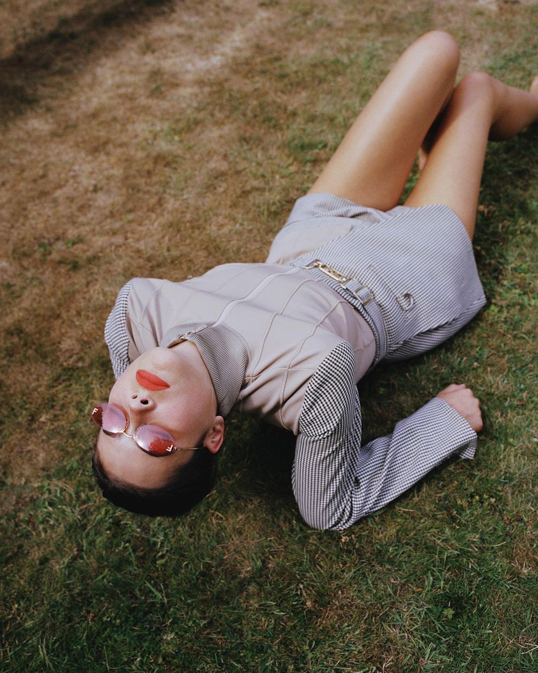 Woman lies on grass wearing striped shorts and blazer with pink sunglasses and lipstick in outdoor setting during daytime.