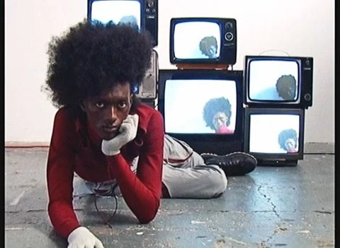 Adult man with afro lying on the floor near a stack of vintage televisions displaying his reflection; indoor scene with neutral background.