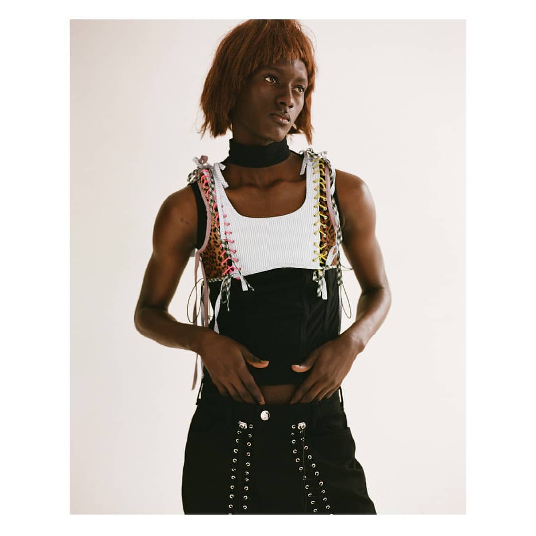 A person poses indoors wearing a black skirt and a top consisting of a white ribbed tank and laced corset.