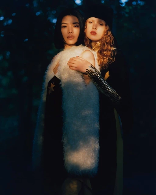 Two women embrace in a dark forest setting. One wears a white faux fur vest, while the other has on a black beret and metallic sleeves.