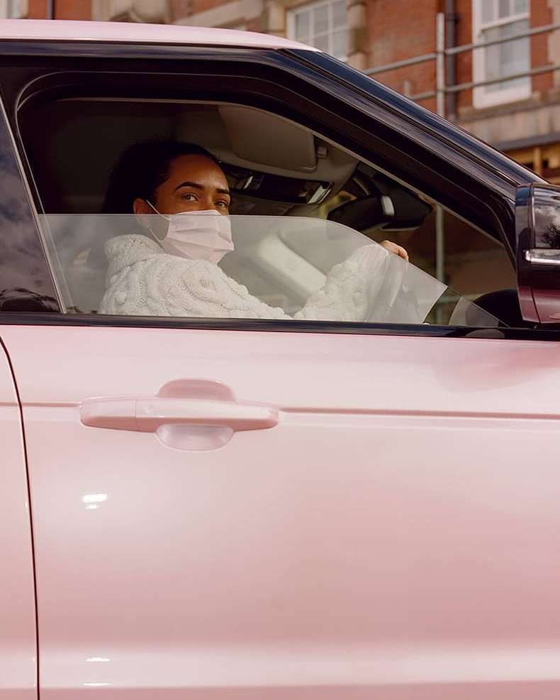 Pink Range Rover parked outdoors, features an adult in the driver's seat wearing a medical face mask and a neutral facial expression.