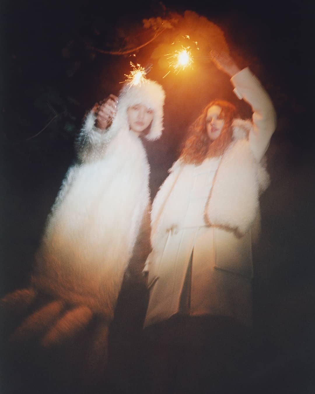 Two people in white furry coats holding sparklers in a dark outdoor setting, creating a festive and surreal fashion moment.