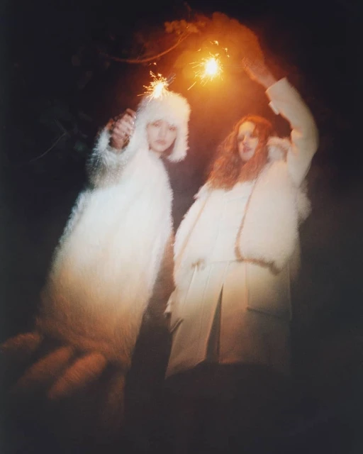 Two people in white furry coats holding sparklers in a dark outdoor setting, creating a festive and surreal fashion moment.
