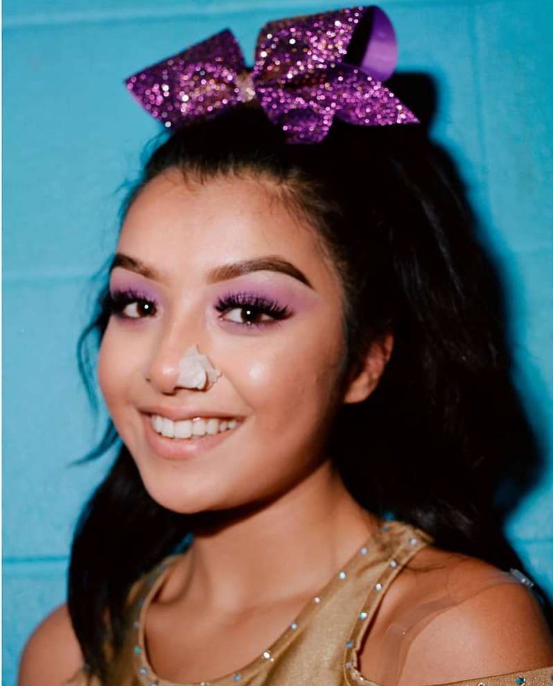 Young woman with purple glitter bow and lavender makeup smiles in a close-up portrait with a light blue background.