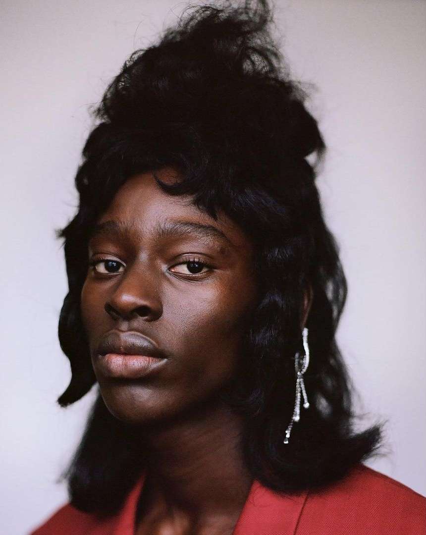 Portrait of a Black woman with dark hair wearing a red jacket and silver earrings against a white background in a close-up shot.