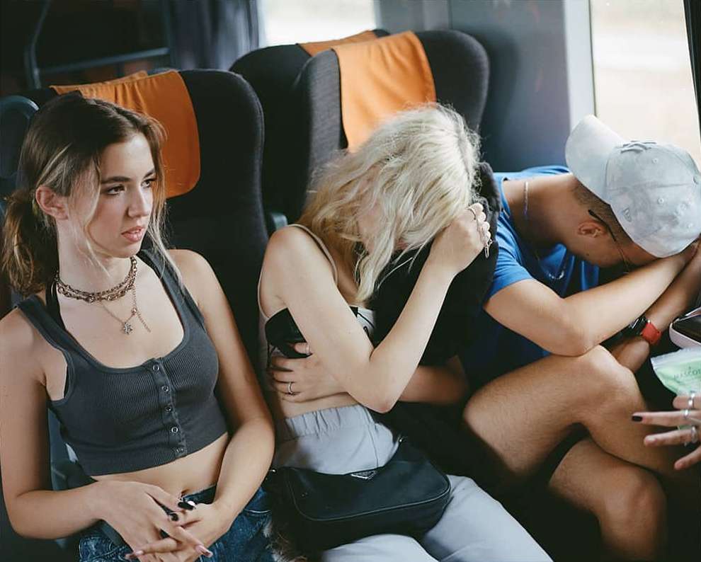 Three people seated together, one with a neutral expression and the others sleeping in a train car with gray and orange seats.