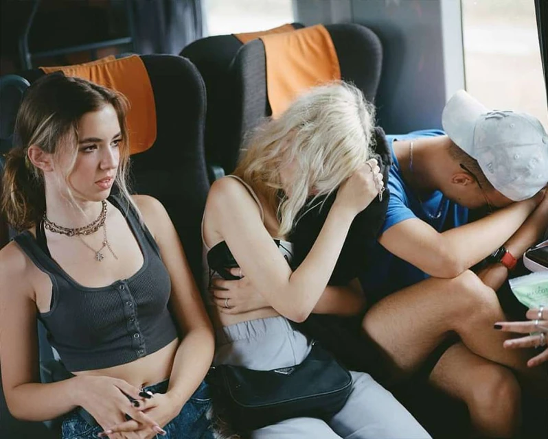 Three people seated together, one with a neutral expression and the others sleeping in a train car with gray and orange seats.