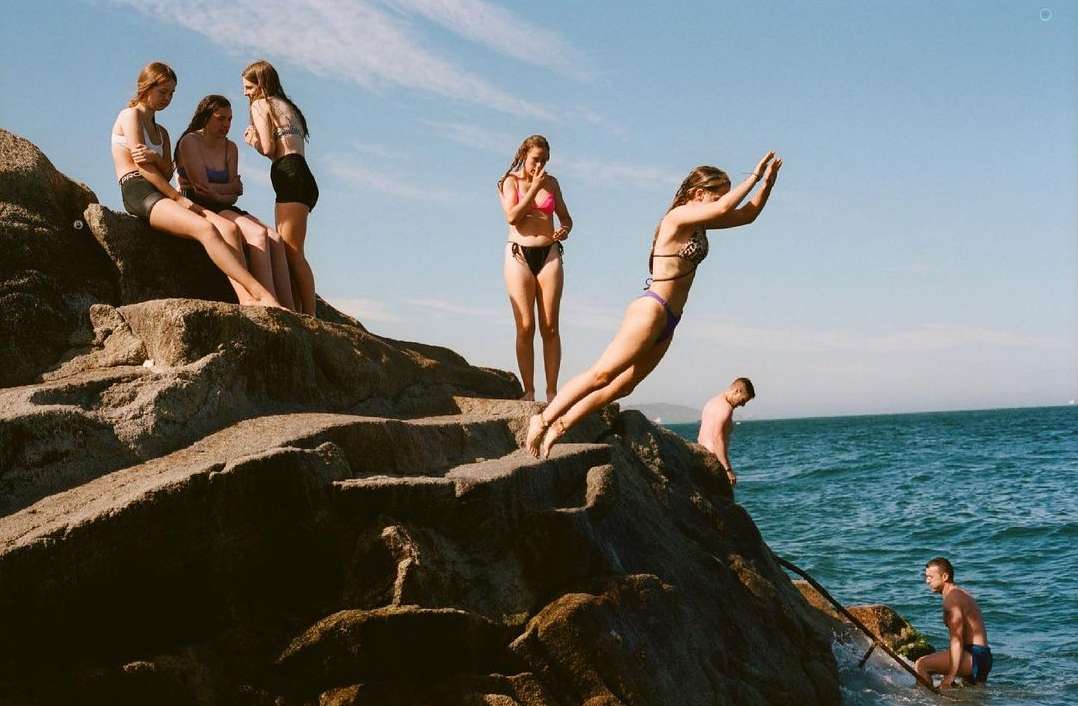 Group of young women and men gather at a rocky beach; one woman dives into the ocean on a sunny day with a clear blue sky above.