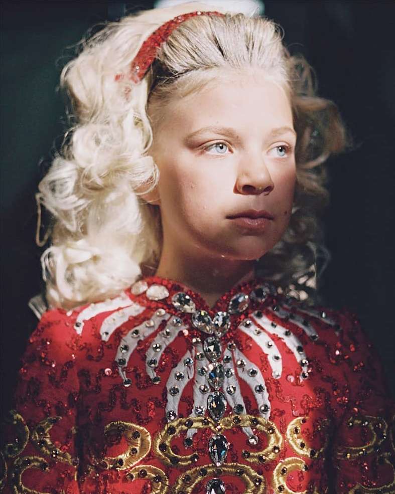 Portrait of a fair-skinned, blue-eyed child with blonde, curly hair. She is wearing a traditional red Irish dance dress with white and gold accents.