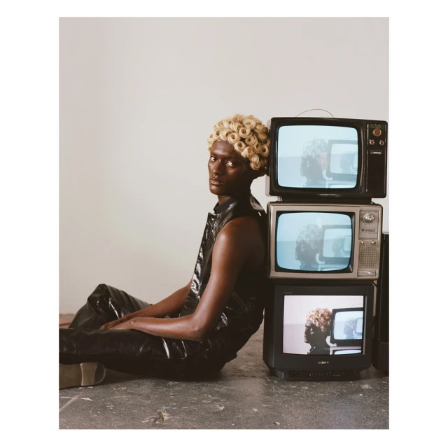 Person with blond wig sitting beside a stack of old televisions, wearing shiny black vest and pants against a neutral background.