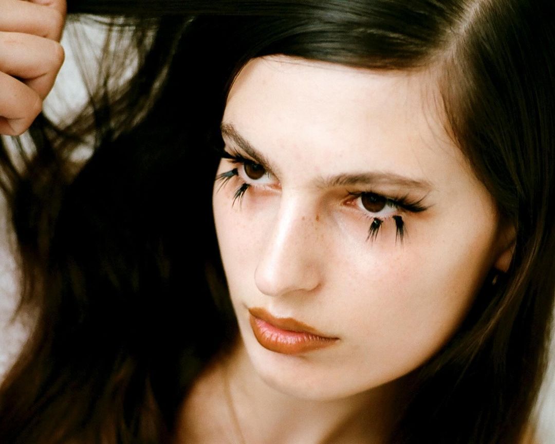 Close-up studio shot of a woman with dark hair and copper lipstick wearing artistic black eyelashes with dark smudges underneath.