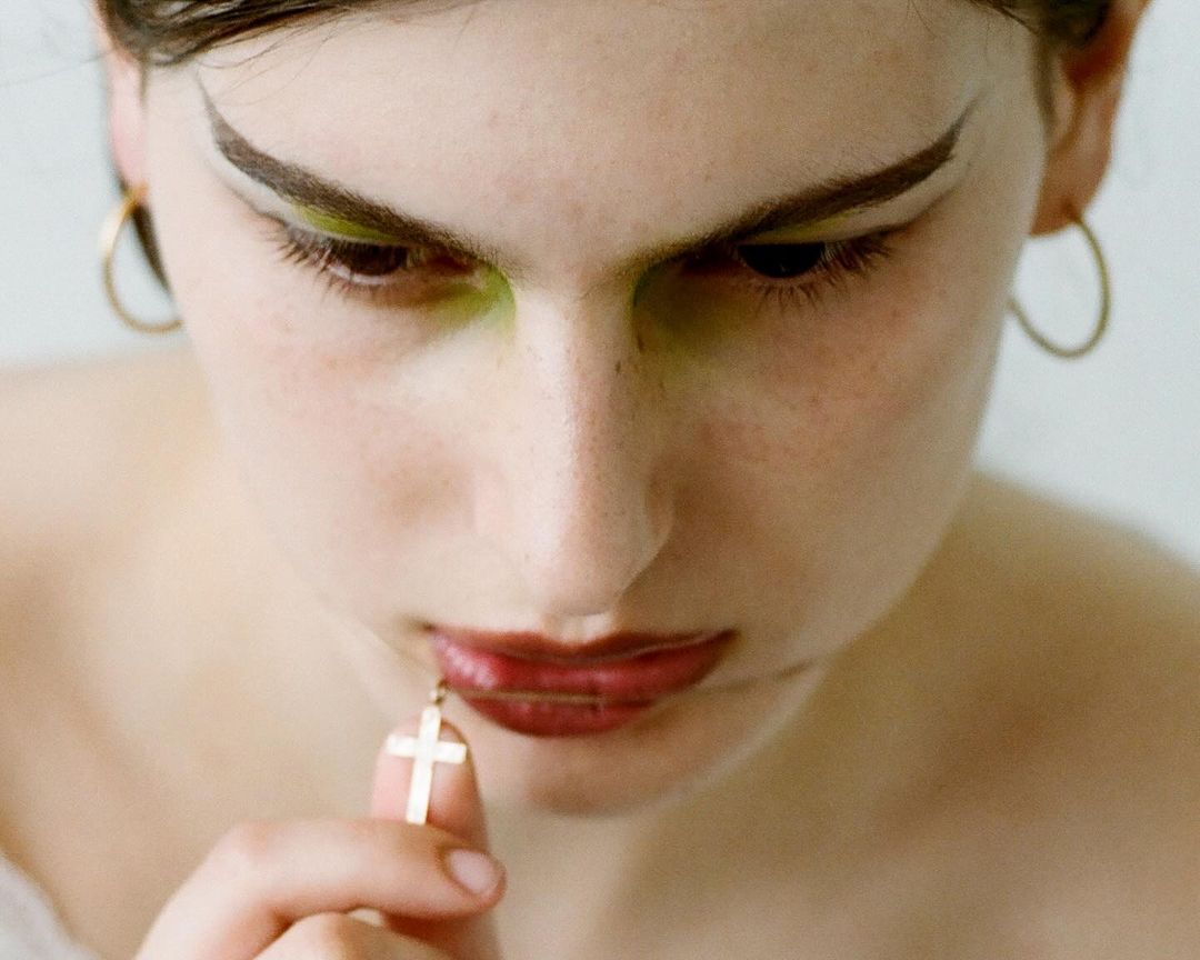 Close-up shot of a woman with green eyeshadow and red lipstick, wearing hoop earrings and holding a gold cross pendant.