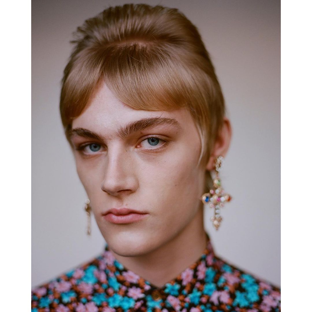 Close-up portrait of a fair-skinned person with blonde hair and blue eyes, wearing colorful floral shirt and ornate cross earrings.