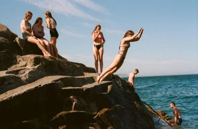 A young woman dives off a rocky coastal ledge into the ocean as friends watch from the cliffs above.