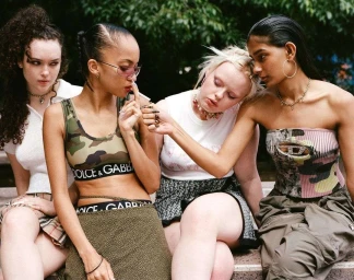 Four diverse young women sitting together outdoors with one woman lighting a cigarette for another in a candid street portrait.