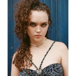 Portrait of a young woman with curly brown hair wearing a patterned top and layered necklace against a blue door