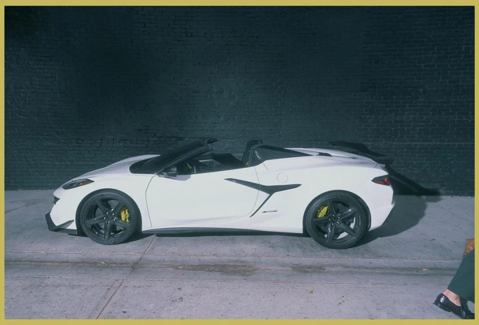 Side profile of a white Chevrolet Corvette convertible with black rims and yellow brake calipers parked against a dark brick wall.