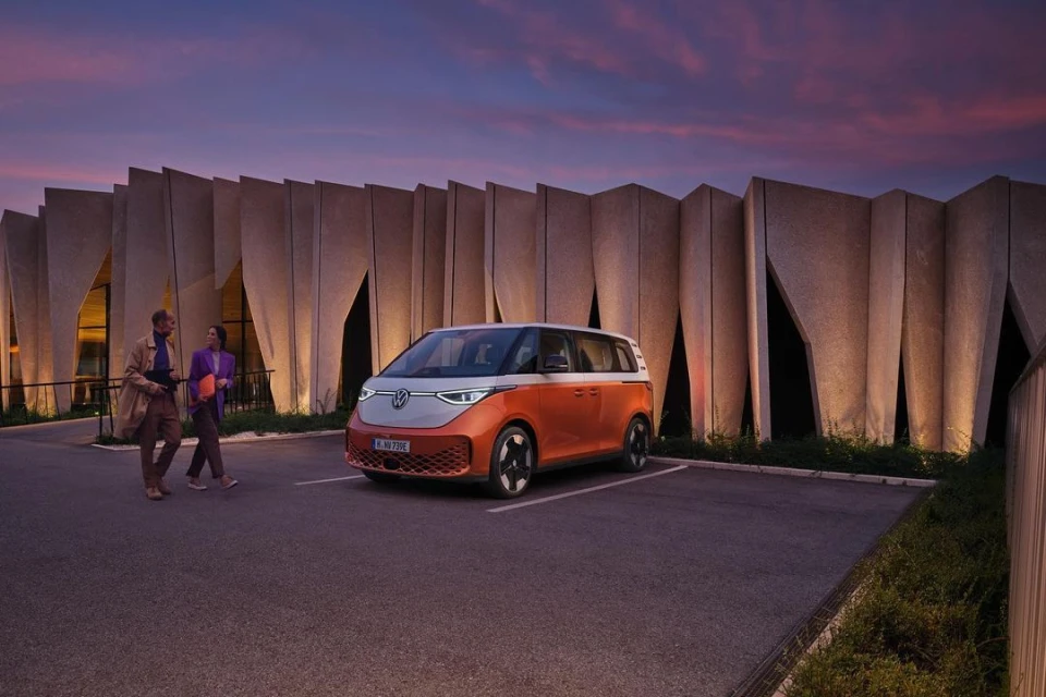 Orange and white VW ID. Buzz electric van parked outdoors with two adults walking nearby in an urban setting with modern architecture.