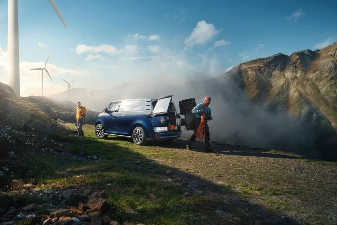 Blue and silver camper van parked on a grassy hillside with two people, mountains, clouds, and wind turbines in the background on sunny day.